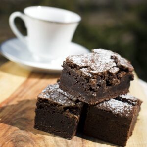Three chocolate brownies on a wooden board in front of a white tea cup